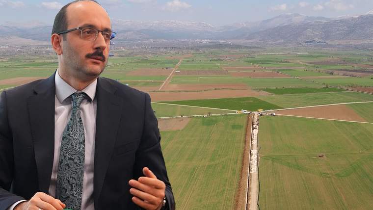 Takım elbiseli ve gözlüklü bir kişi, ekili tarlalar ile sulama kanalı görülen manzaranın önünde duruyor.