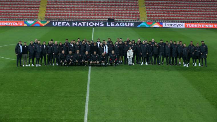 Futbol sahasında, kırmızı tribünlerin önünde yan yana duran kalabalık bir takım ve teknik ekip görülüyor.