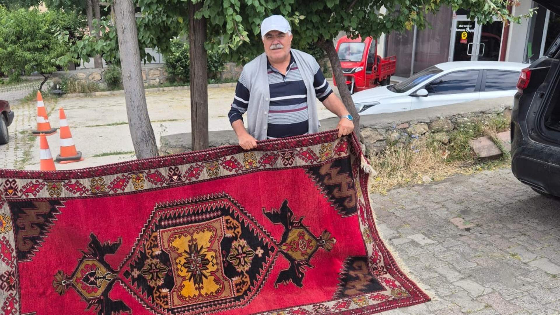Bir adam, elinde büyük bir halı tutuyor. Arka planda park halindeki araçlar ve ağaçlar görünmekte.