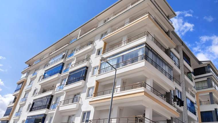 Balkonları ve camla kaplı bölümleri bulunan, çok katlı beyaz bir apartman binası görülüyor.