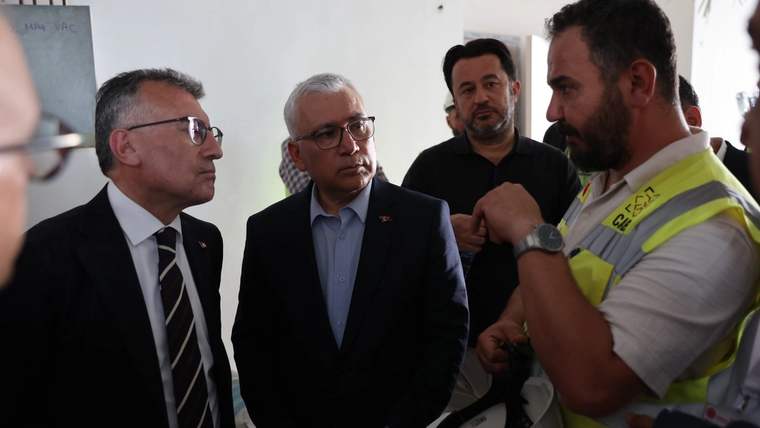 İnşaat alanında, iki kişi bir inşaat görevlisiyle konuşuyor. Arka planda diğer kişiler ve inşaat malzemeleri görünüyor.