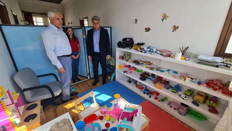 Bir odada üç kişi, oyuncaklarla dolu rafların ve çocuk etkinlik masasının yanında duruyor.