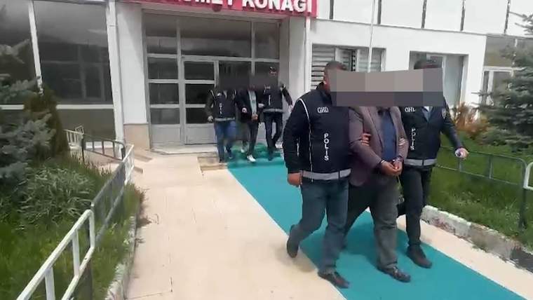 Yüzleri bulanık iki kişi, polis yelekli görevliler eşliğinde bir binanın girişinden çıkıyor.