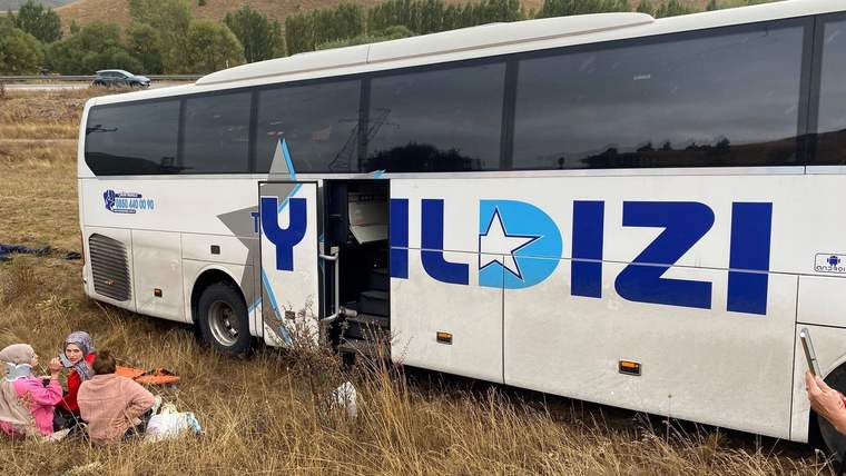 Çalılık alanda duran beyaz bir yolcu otobüsünün kapısı açık; yanında yerde oturan kişiler görülüyor.