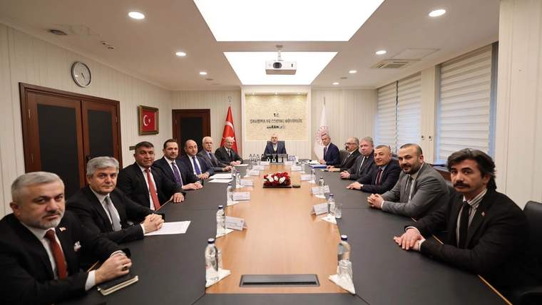 Toplantı odasında uzun masa çevresinde oturan kişiler, Türk bayrağı ve duvarda asılı bir portre görülüyor.