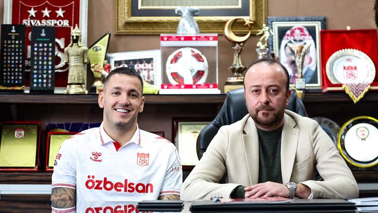 Sivasspor forması giymiş bir futbolcu, masada oturan bir kişiyle birlikte poz veriyor.