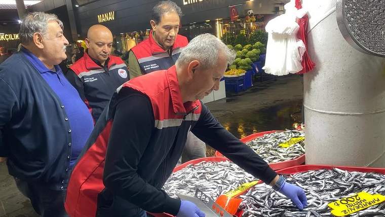 Balık tezgâhındaki balıkları eldivenli elleriyle inceleyen bir görevli ve yanında duran üç kişi görülüyor.