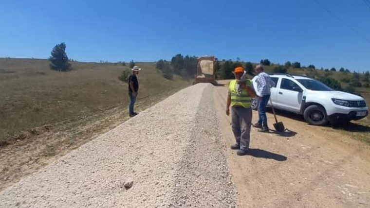 Kırsal alanda stabilize yol üzerinde kamyon ve üç kişi, yol kenarında beyaz bir araçla görülüyor.