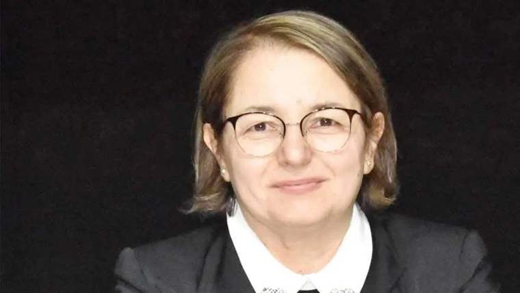 Kısa saçlı, gözlüklü bir kadın siyah ceket ve beyaz yakalı bluzla kameraya bakıyor.