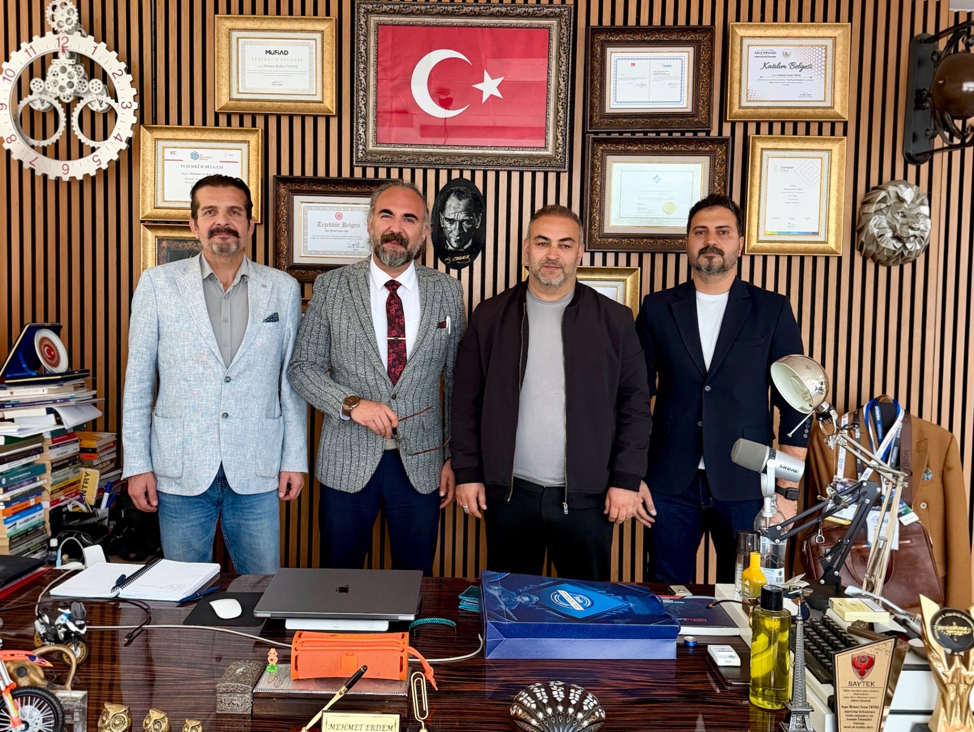 Duvarında Türk bayrağı ve çok sayıda çerçeve bulunan ofiste, dört kişi masa arkasında yan yana duruyor.