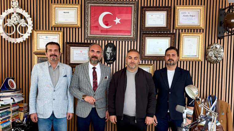 Duvarında Türk bayrağı ve çok sayıda çerçeve bulunan ofiste, dört kişi masa arkasında yan yana duruyor.