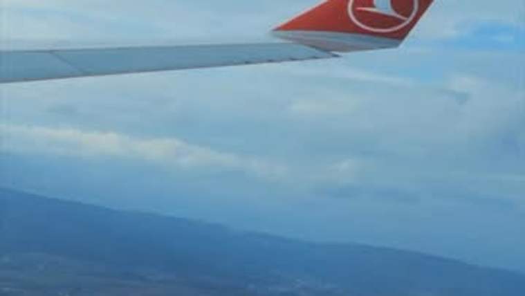 Uçak kanadının görünümü, arka planda bulutlu bir gökyüzü ve dağlık alanlar yer alıyor.