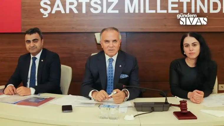 Toplantı masasında oturan üç kişi, önlerindeki belgeler, mikrofonlar ve kırmızı tokmakla birlikte görülüyor.