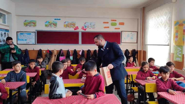 Sınıfta, masalarda oturan çocuklara yaklaşan bir adam, elinde bir paket tutuyor.