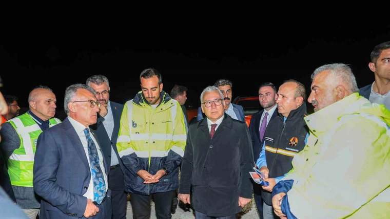 Gece karanlığında, takım elbiseli ve reflektif yelekli kişiler asfalt alanda birlikte duruyor.