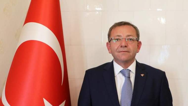 Türk bayrağının yanında koyu renk takım elbiseli ve kravatlı bir kişi, kameraya doğru duruyor.
