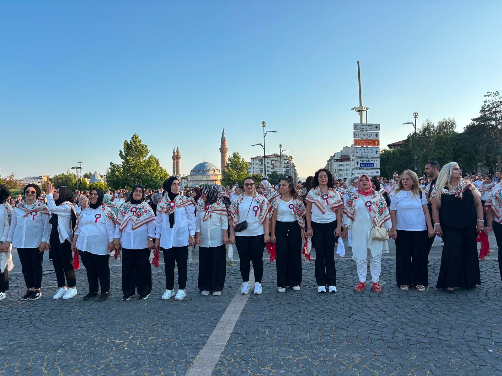 Kaldırımda yan yana duran, beyaz giysili ve kırmızı desenli şallar takan kalabalık grup