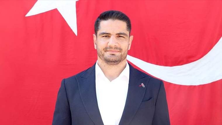 Beyaz gömlek ve siyah ceket giymiş bir kişi, arkasında Türk bayrağı ile poz veriyor.
