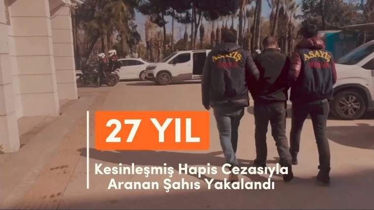 Üzerlerinde “ASAYİŞ” yazılı yelekler bulunan iki kişi, ortalarındaki bir adamla birlikte açık alanda yürüyor.