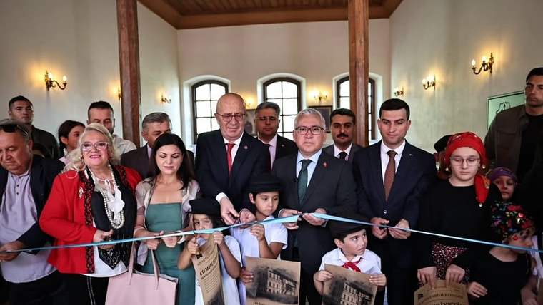 İç mekânda yetişkinler ve çocuklar, ellerindeki makaslarla mavi kurdeleyi keserken görülüyor.