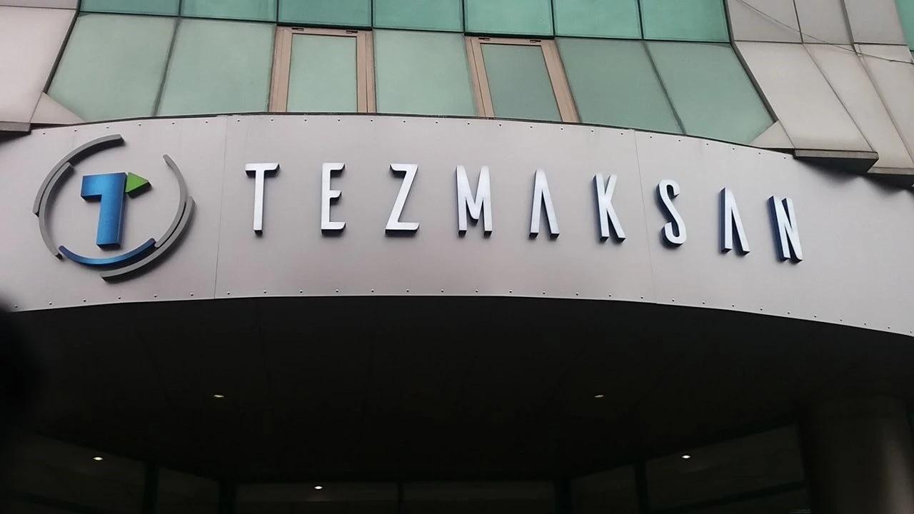Bir binanın girişinde, üzerinde “TEZMAKSAN” yazısı ve mavi-turuncu logonun bulunduğu tabela görülüyor.