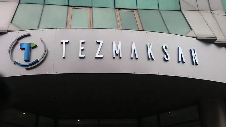 Bir binanın girişinde, üzerinde “TEZMAKSAN” yazısı ve mavi-turuncu logonun bulunduğu tabela görülüyor.