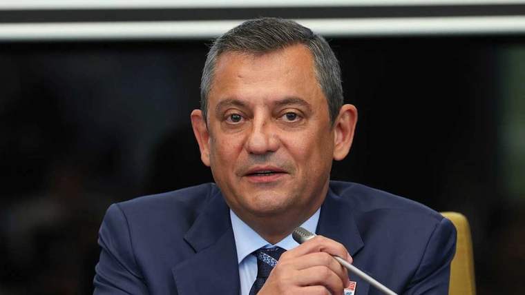 Koyu renk takım elbiseli bir kişi, elindeki mikrofonla konuşurken kameraya bakıyor.