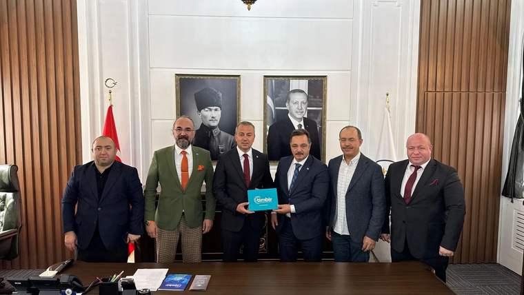 Bir odada altı erkek yan yana duruyor; ortadaki kişi üzerinde “TİMBİR” yazan turkuaz plaketi tutuyor.