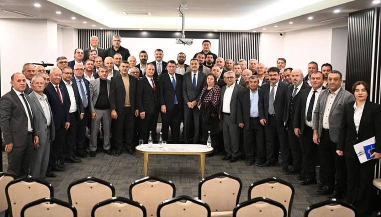 Toplantı salonunda, takım elbiseli çok sayıda kişi kamera karşısında yan yana duruyor; önde masa ve sandalyeler var.
