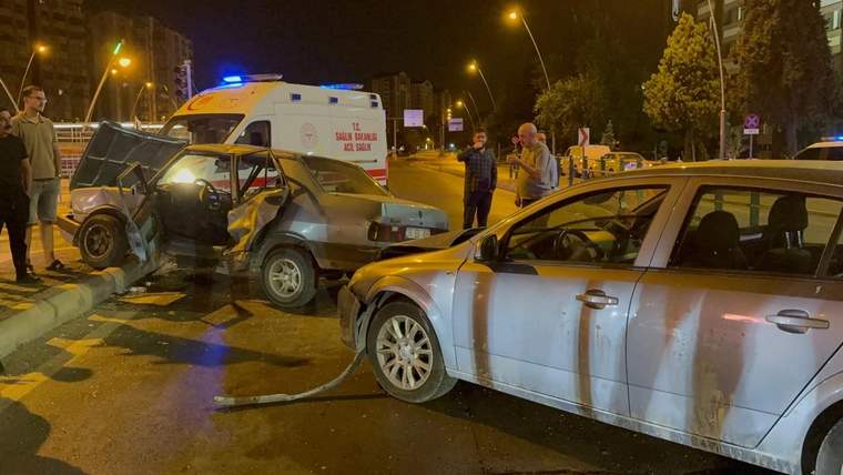 Gece aydınlatılmış yolda hasarlı iki otomobil, bir ambulans ve araçların yanında duran iki kişi görülüyor.