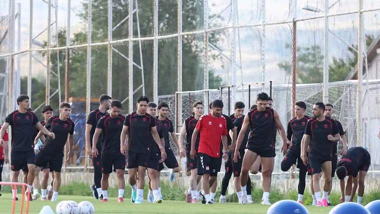 Antrenman sahasında siyah ve kırmızı formalar giyen bir grup futbolcu, toplar ve koniler arasında yürüyor.