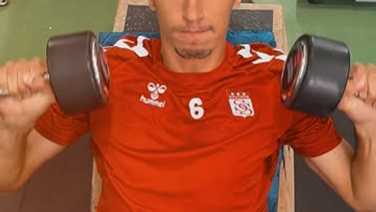 Kırmızı forma giymiş bir sporcu, dambıl kaldırarak antrenman yapıyor.