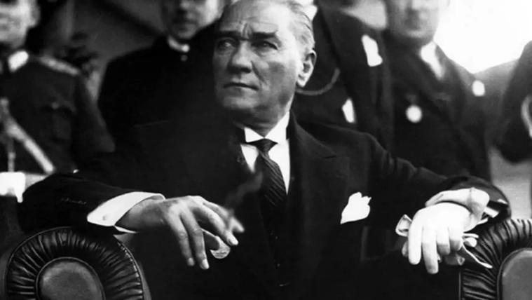 Atatürk, bir koltukta otururken, elinde sigara tutuyor ve ciddi bir ifadeye sahip.
