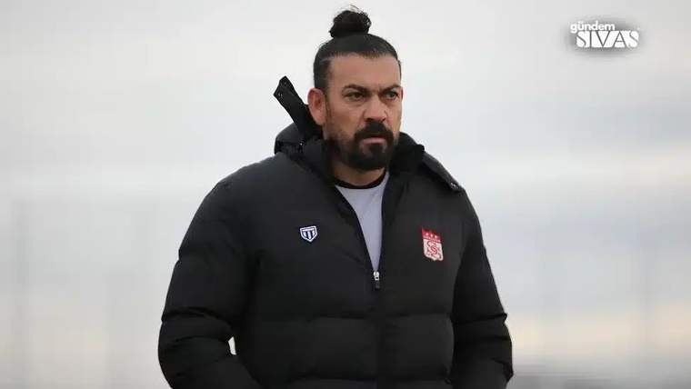 Sakallı bir kişi, siyah montuyla açık havada ayakta duruyor; montunda iki arma bulunuyor.