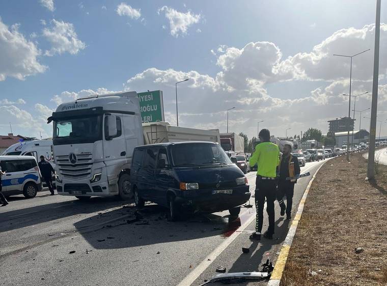 Yol üzerinde duran beyaz kamyon ve siyah minibüsün çevresinde iki kişi, araç parçaları ve yoğun trafik görülüyor.