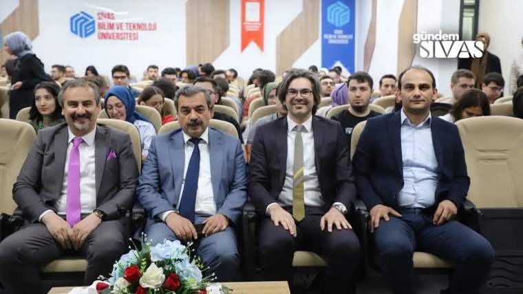 Bir salonda dört takım elbiseli kişi önde oturuyor; arkalarında izleyiciler ve üniversite afişleri görülüyor.