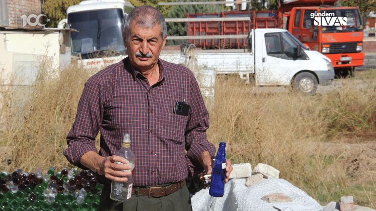 Bir adam, elinde bir şişe tutarak arka planda şişe yığını ve araçlar ile birlikte duruyor.