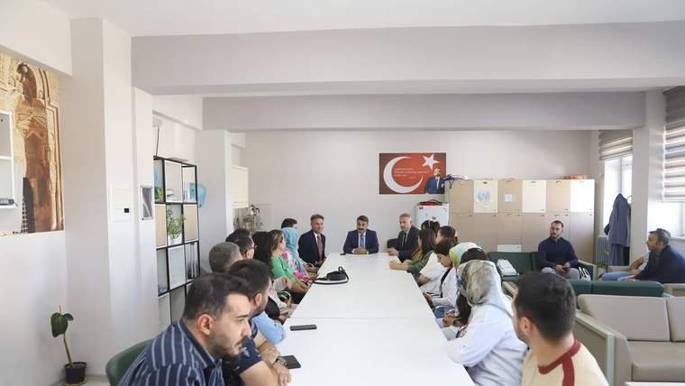 Bir odada uzun masa çevresinde oturan yetişkinler ve öğrenciler, karşılıklı görüşme yapıyor; duvarda Türk bayrağı var.