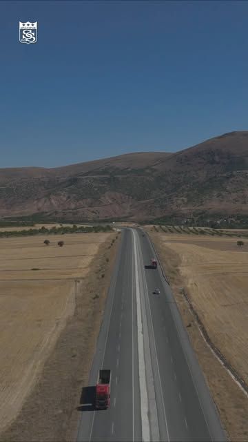 Kurak tarlalar ve dağlar arasından uzanan bölünmüş yolda birkaç araç ilerliyor.