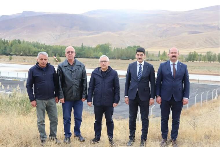 Kurak arazide, arkalarında tel çit ve ağaçlı tepeler bulunan yan yana duran beş kişi
