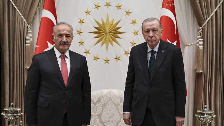 Takım elbiseli iki kişi, Türk bayrakları ve yıldızlarla çevrili altın renkli güneş motifinin önünde duruyor.