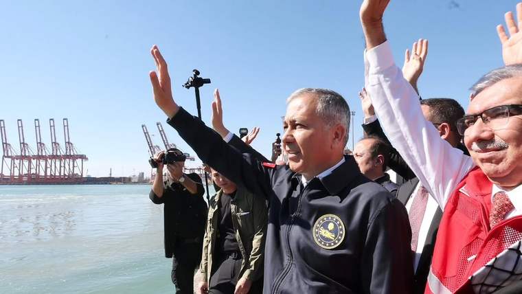 Liman kıyısında, üzerinde arma bulunan yelekli bir kişiyle birlikte birkaç kişi el sallıyor; vinçler görülüyor.