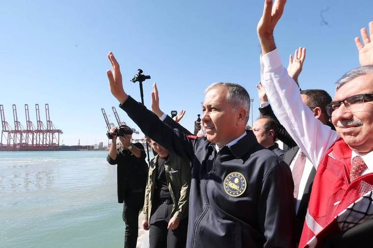 Liman kıyısında, üzerinde arma bulunan yelekli bir kişiyle birlikte birkaç kişi el sallıyor; vinçler görülüyor.