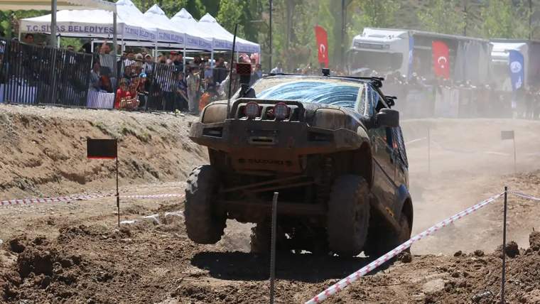 Zorlu parkurda havada bir off-road aracı, etrafında kalabalık izleyiciler var.