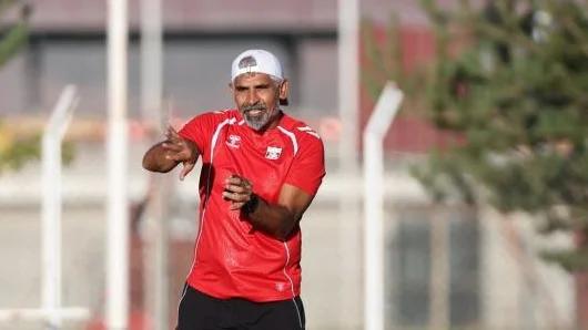 Kırmızı forma giymiş bir antrenör, antrenman sahasında oyunculara talimat veriyor.
