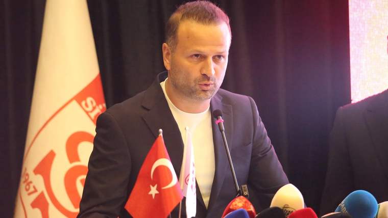 Mikrofonlar önünde konuşan bir erkek, yanında Türk bayrağı ve kırmızı-beyaz flamalar bulunuyor.