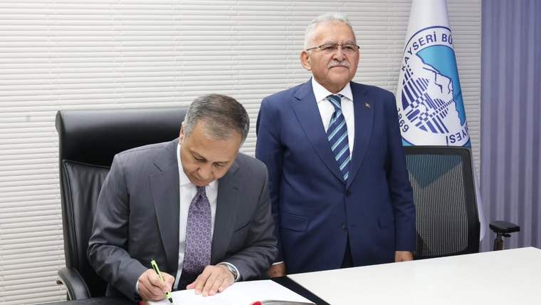 Bir kişi masada kırmızı kaplı deftere yazarken, arkasında duran takım elbiseli kişi ve Türk bayrağı görülüyor.