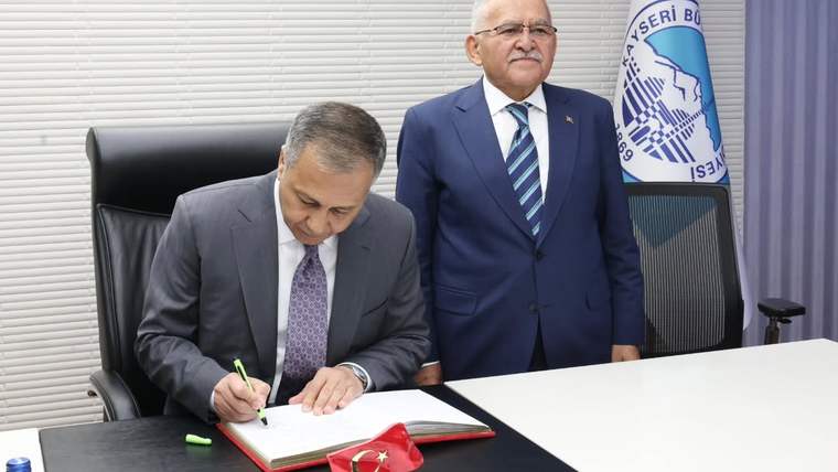 Bir kişi masada kırmızı kaplı deftere yazarken, arkasında duran takım elbiseli kişi ve Türk bayrağı görülüyor.
