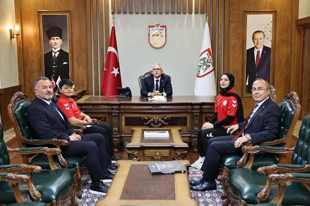 Bayraklar ve portrelerin bulunduğu makam odasında, masa çevresinde oturan beş kişi görülüyor.
