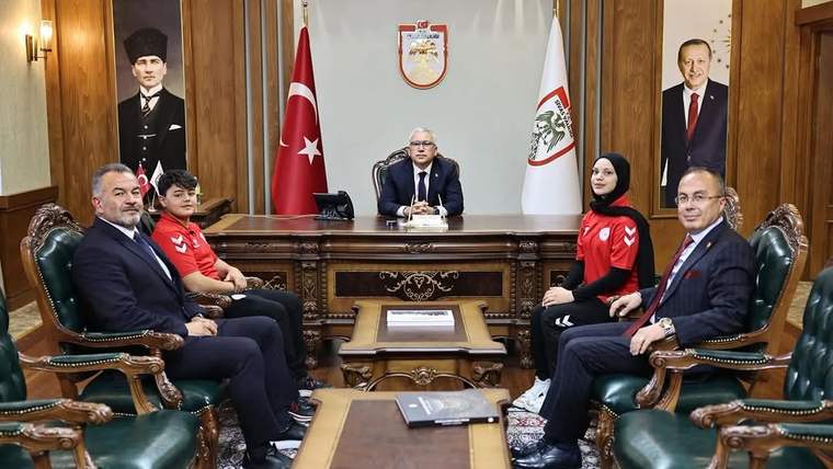 Bayraklar ve portrelerin bulunduğu makam odasında, masa çevresinde oturan beş kişi görülüyor.